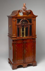 Mahogany cabinet with a glass panel revealing brass tubes and a carved bust on top.