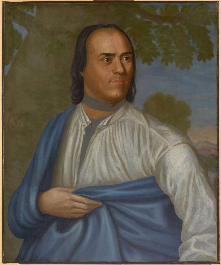 Portrait of a Native American man with long black hair, draped in a blue sheet and posed in front of a tree with blue sky in the background.