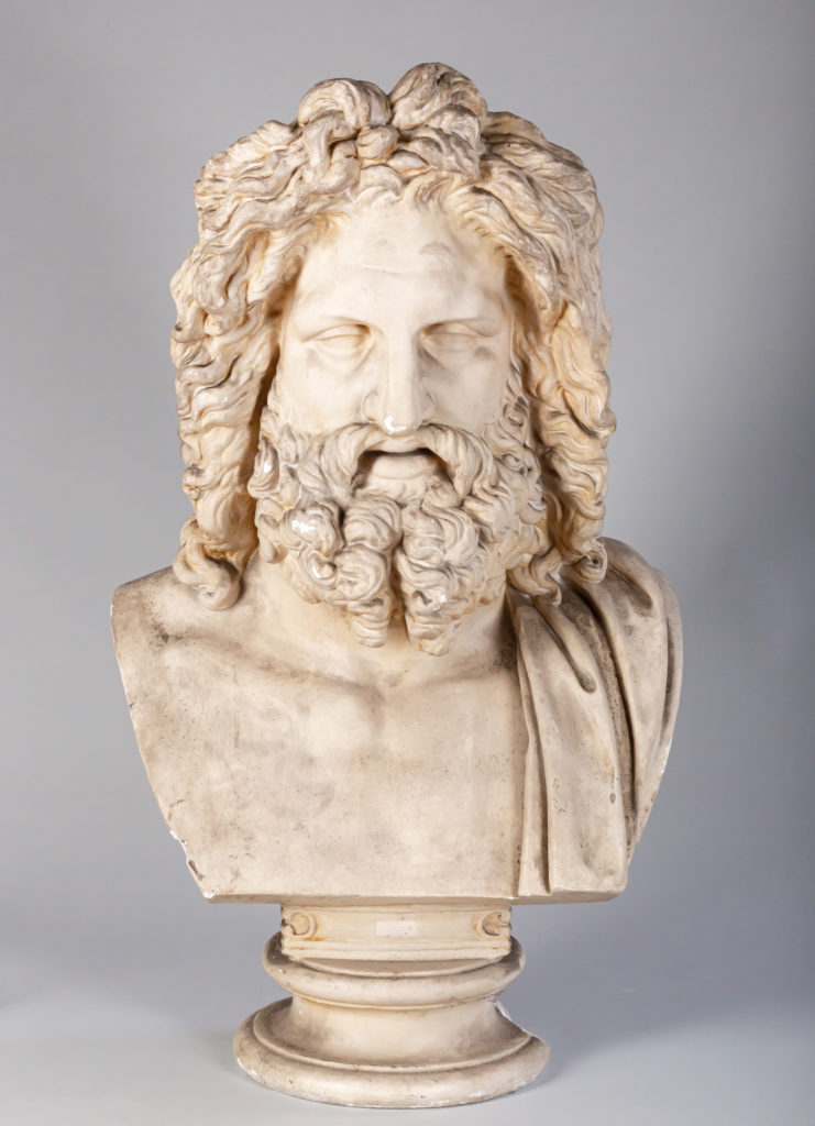 Plaster bust of a man with long curly hair and a long beard, fabric draped over his left shoulder.