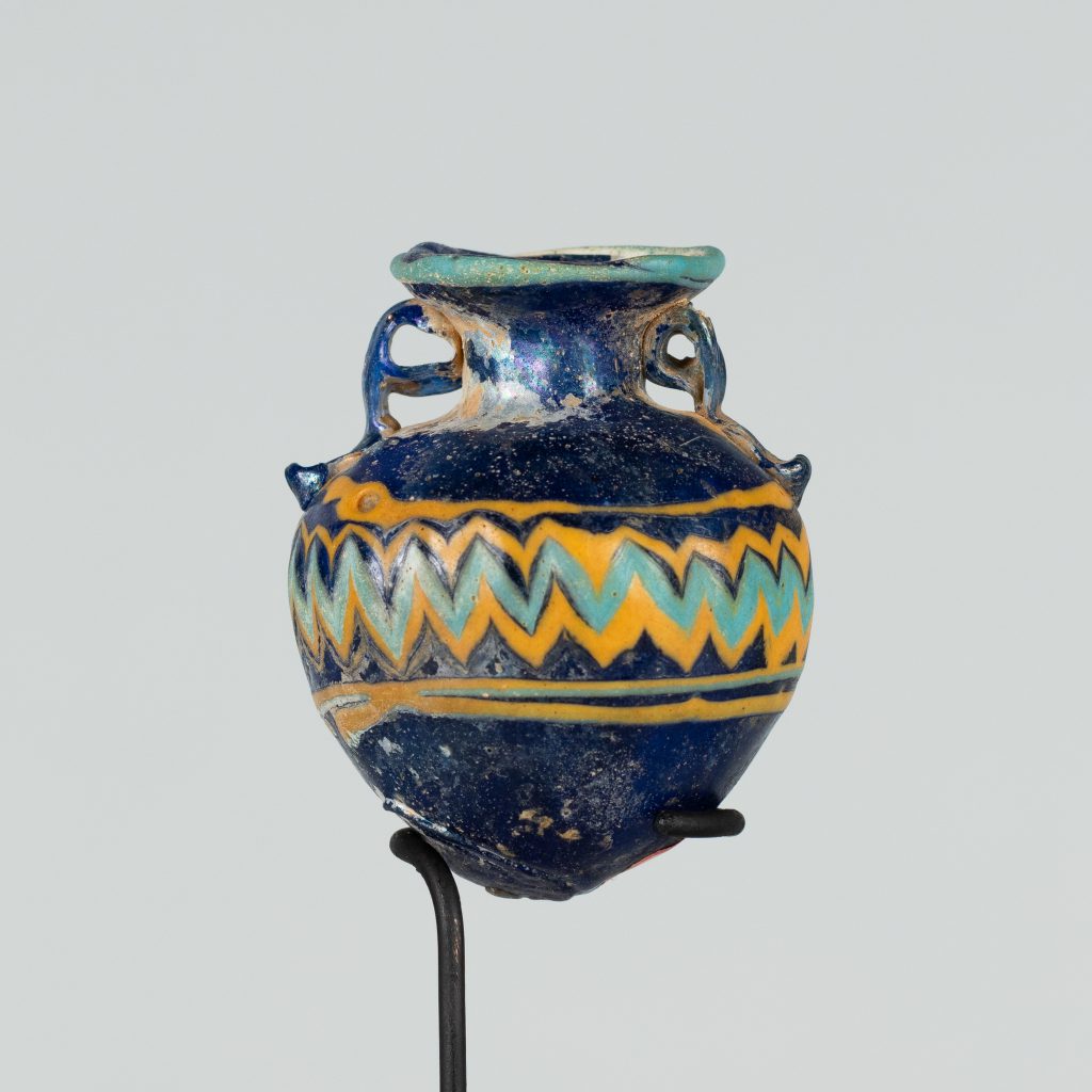 An ancient roundish glass vessel with two small handles and a short narrower neck opening to a wider mouth. It is painted navy blue and has yellow and turquoize stripes and a chevron pattern. The lip is painted turquoise.