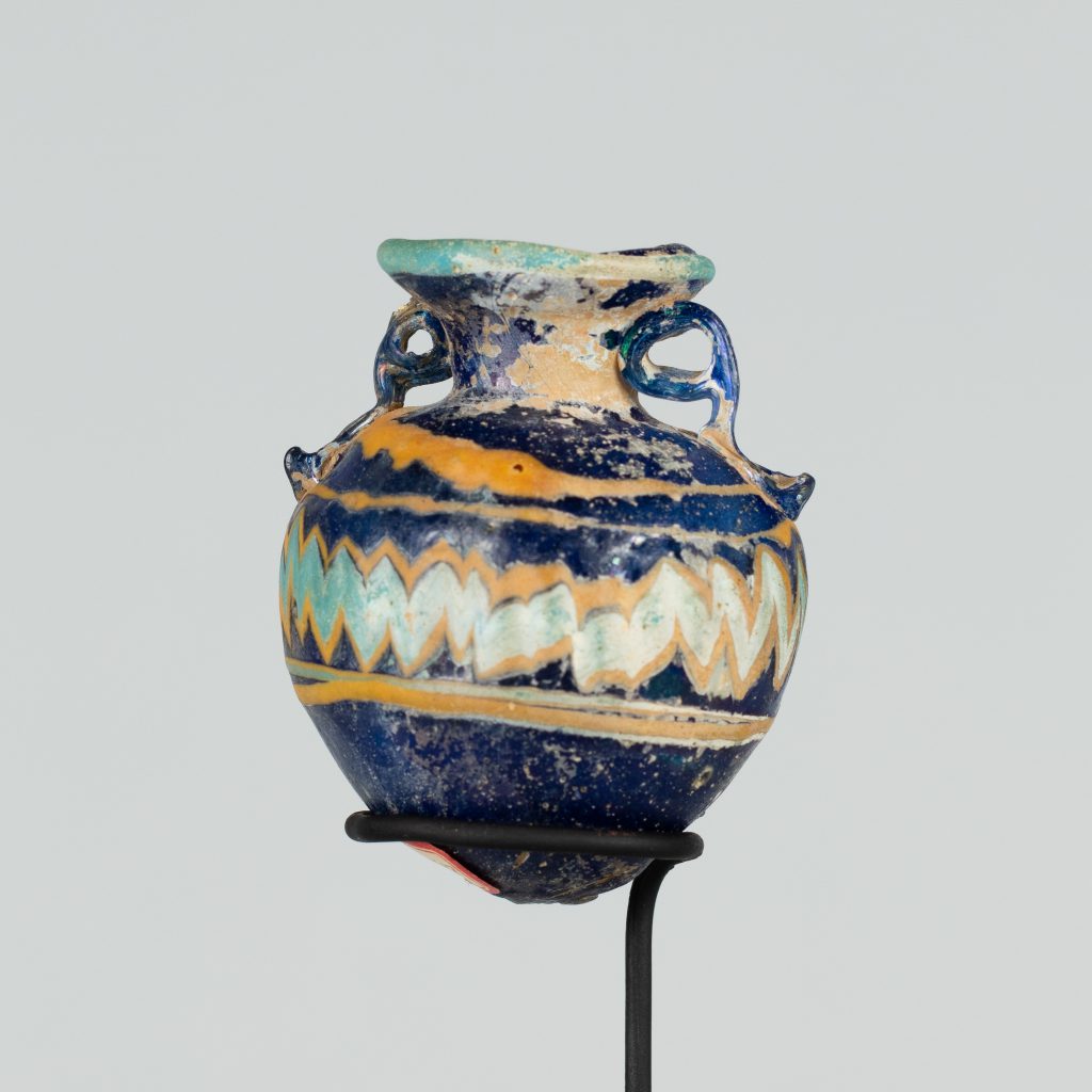 Alternate view of An ancient roundish glass vessel with two small handles and a short narrower neck opening to a wider mouth. It is painted navy blue and has yellow and turquoize stripes and a chevron pattern. The lip is painted turquoise.