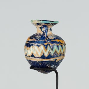 Alternate view of An ancient roundish glass vessel with two small handles and a short narrower neck opening to a wider mouth. It is painted navy blue and has yellow and turquoize stripes and a chevron pattern. The lip is painted turquoise.
