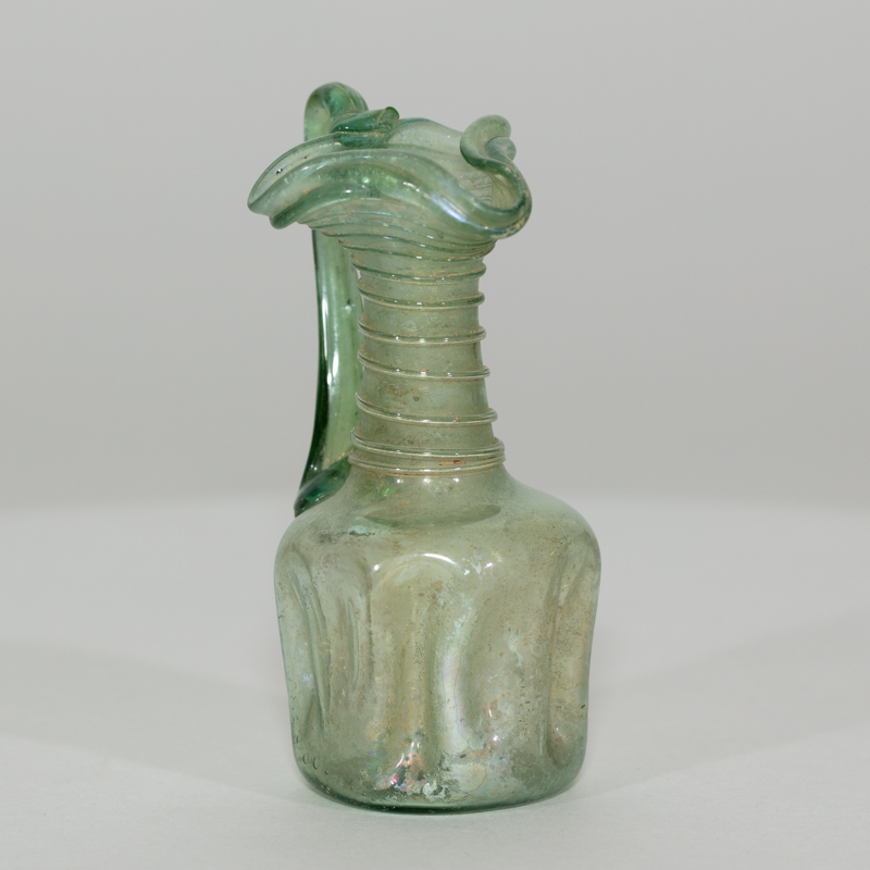 Alternate view of An ancient minature, one-handled, ornately-decorated, glass jug tinted green. Its neck is decorated with glass rings and its lip is wavy.