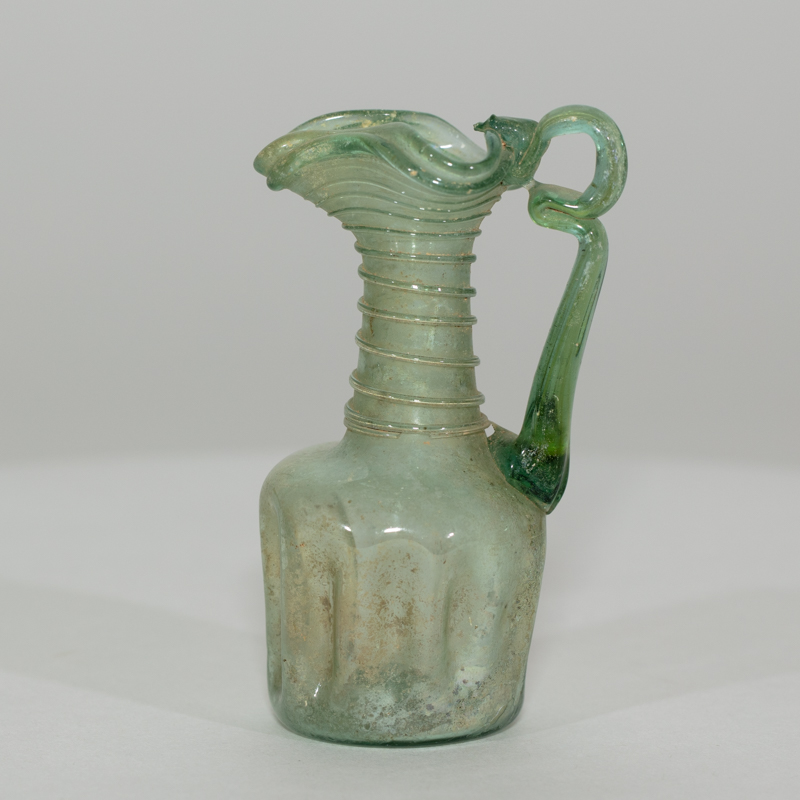 Alternate view of An ancient minature, one-handled, ornately-decorated, glass jug tinted green. Its neck is decorated with glass rings and its lip is wavy.