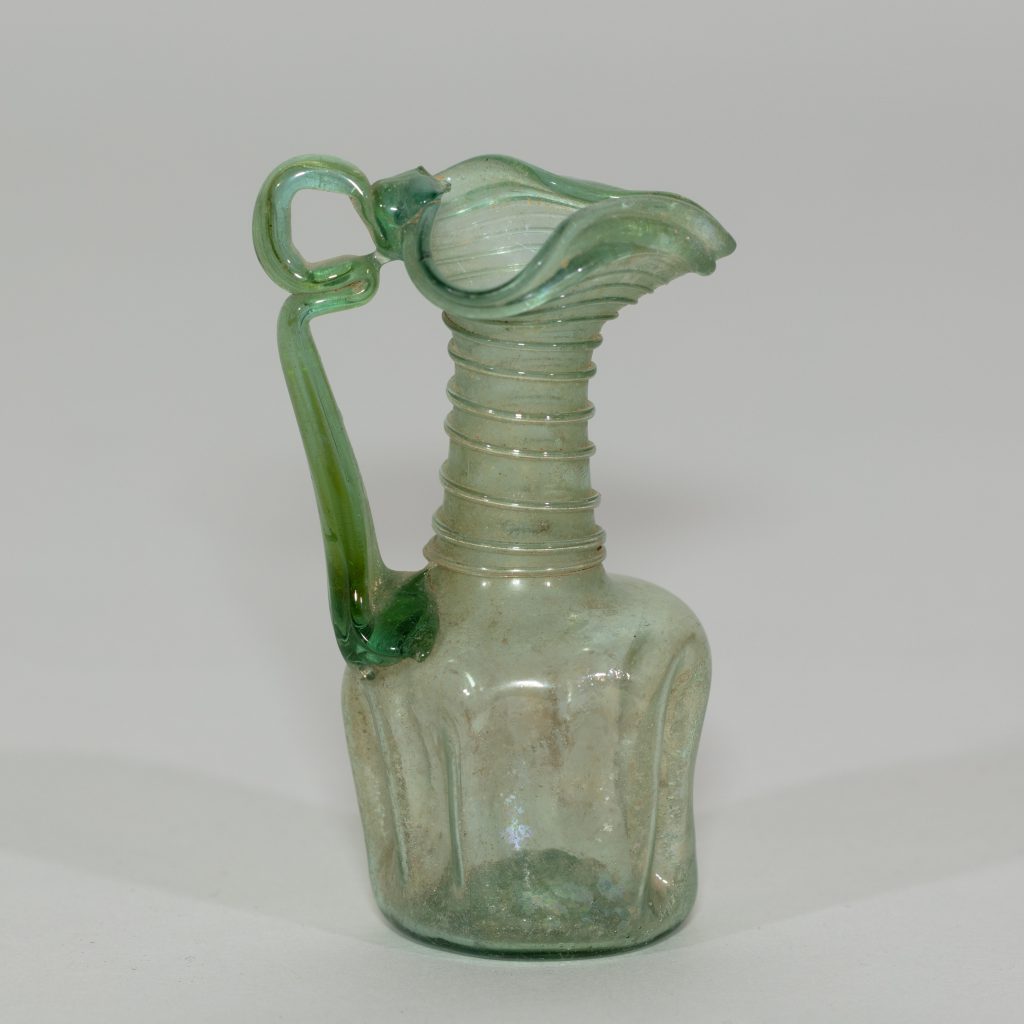 An ancient minature, one-handled, ornately-decorated, glass jug tinted green. Its neck is decorated with glass rings and its lip is wavy.