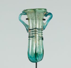 An ancient narrow, two-handled glass vessel tinted teal. The vessel is a long pipe shape folded in half so the two opening meet at the top.