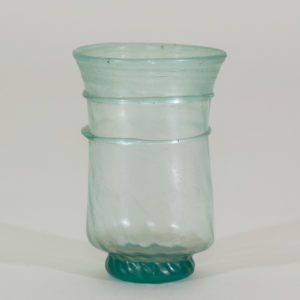 An ancient small, transparent, handless, glass cup tinted teal. It has 2 glass 2 perimeter rings and a narrower solid teal base.