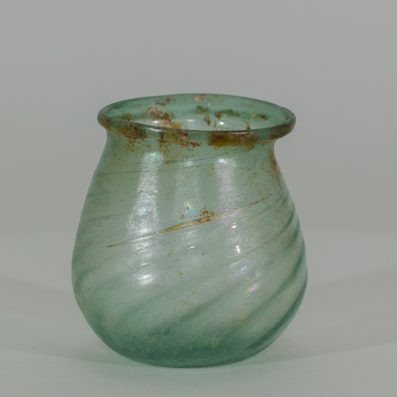 Alternate view of An ancient small, paritally-transparent glass jar tinted green with some brownish paint marks toward the lip.