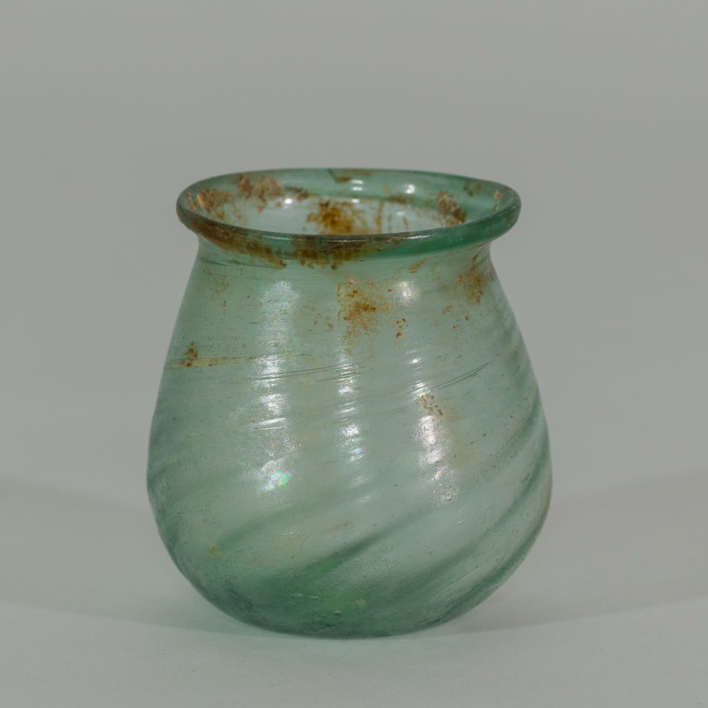 An ancient small, paritally-transparent glass jar tinted green with some brownish paint marks toward the lip.