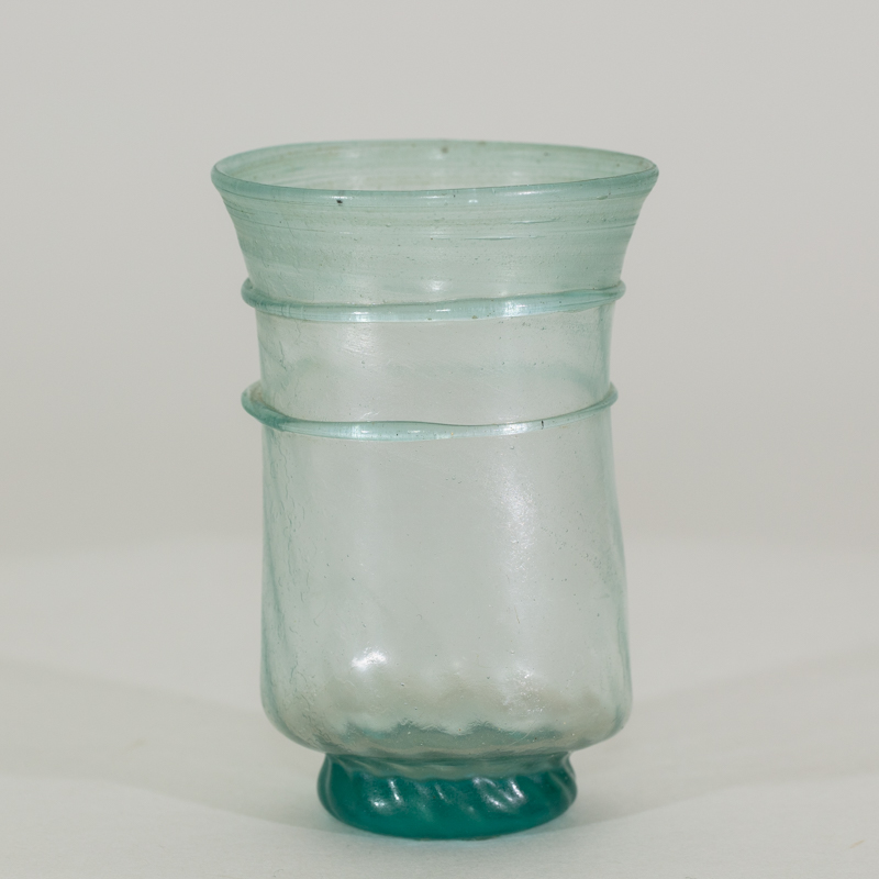 Alternate view of An ancient small, transparent, handless, glass cup tinted teal. It has 2 glass 2 perimeter rings and a narrower solid teal base.