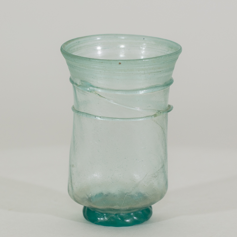 Alternate view of An ancient small, transparent, handless, glass cup tinted teal. It has 2 glass 2 perimeter rings and a narrower solid teal base.