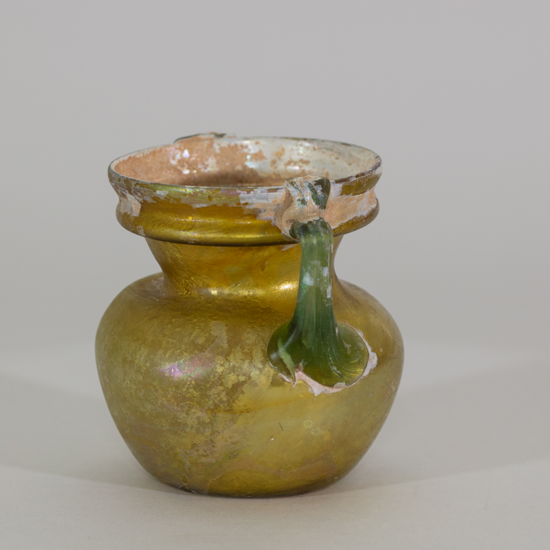 Alternate view of An ancient two-handled glass vessel with green handles, a pearl-colored base, that becomes gold moving up the vessel.
