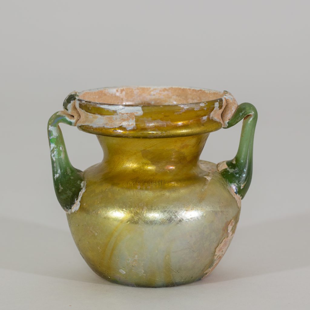 An ancient two-handled glass vessel with green handles, a pearl-colored base, that becomes gold moving up the vessel.