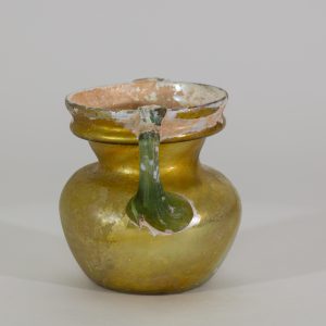 Alternate view of An ancient two-handled glass vessel with green handles, a pearl-colored base, that becomes gold moving up the vessel.