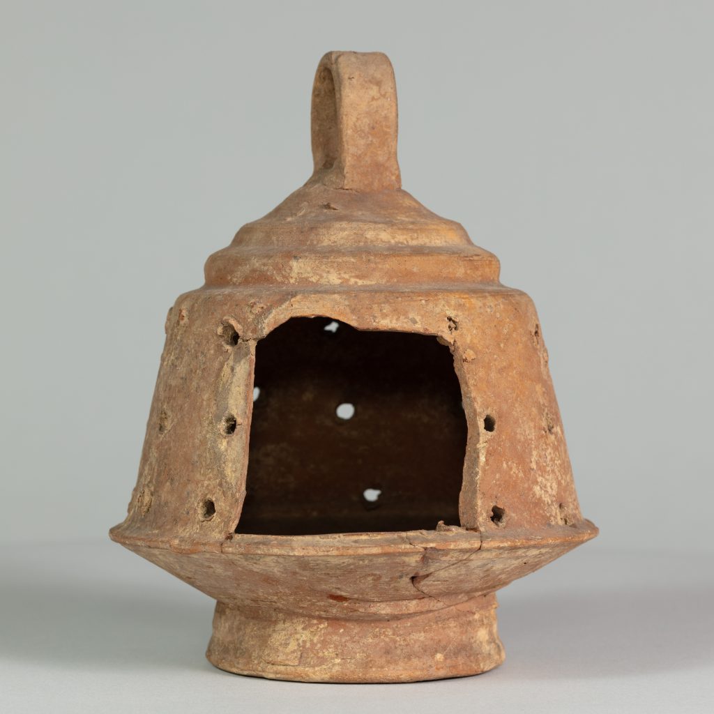 Alternate view of A ancient, ceramic, rust-colored, bell-shaped lantern with an opening on one side, a top handle, and holes perferated around the perimeter.