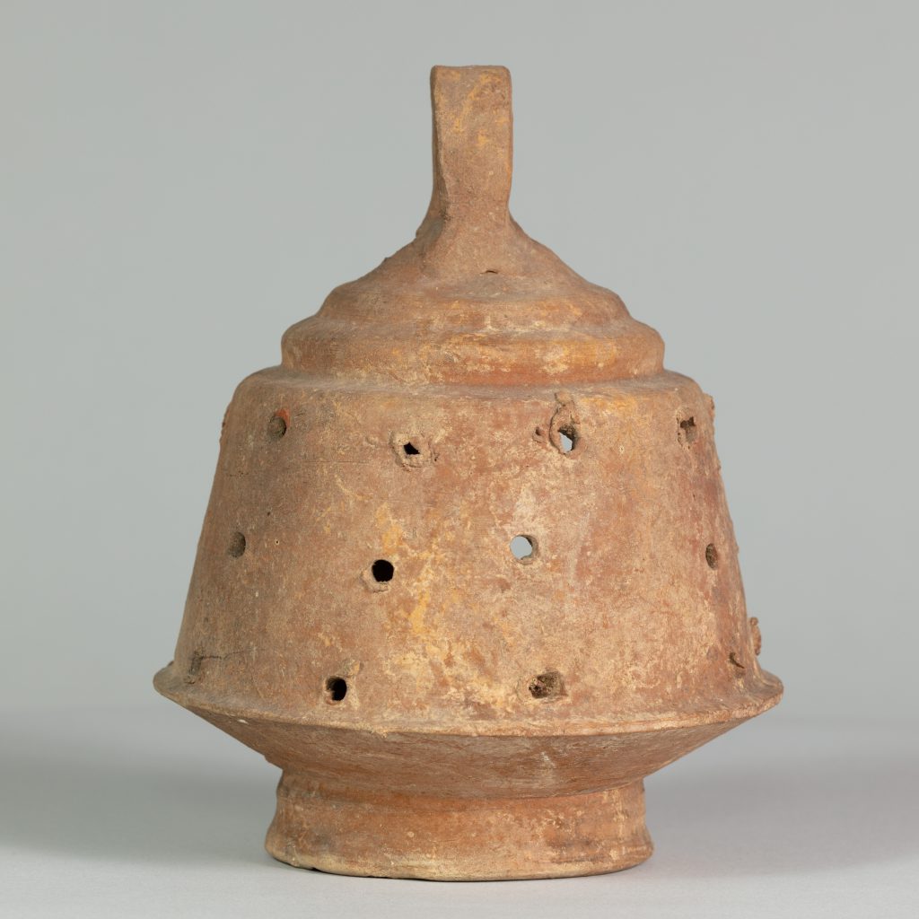 Alternate view of A ancient, ceramic, rust-colored, bell-shaped lantern with an opening on one side, a top handle, and holes perferated around the perimeter.