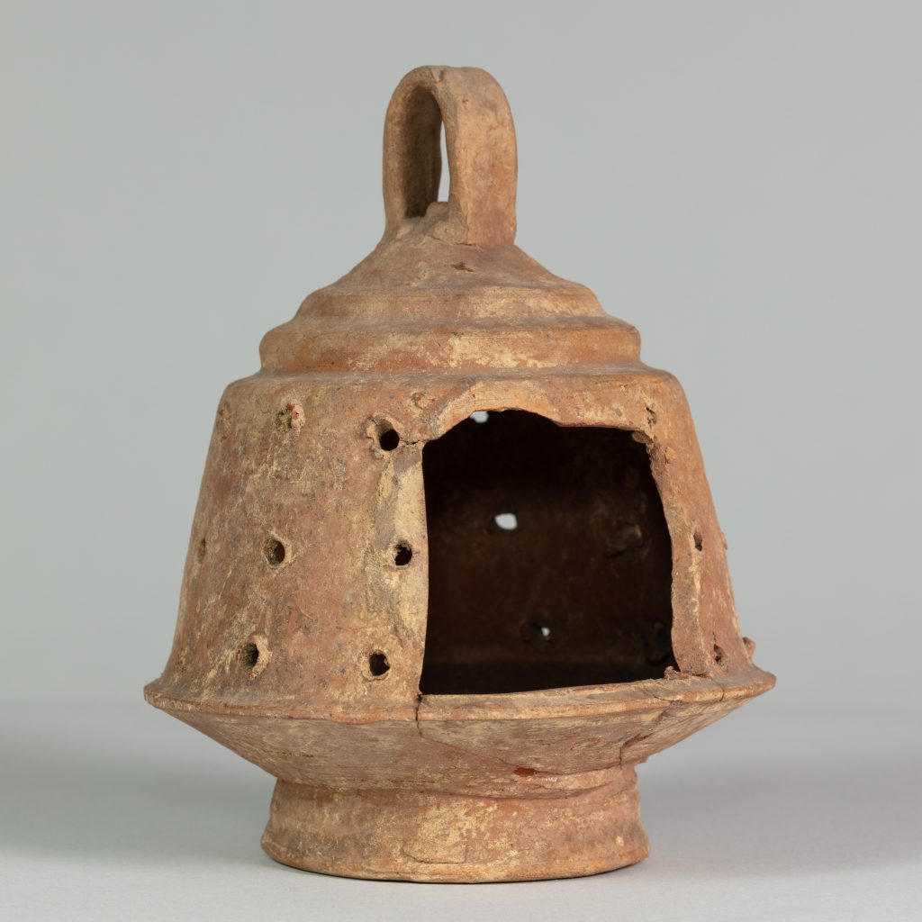 A ancient, ceramic, rust-colored, bell-shaped lantern with an opening on one side, a top handle, and holes perferated around the perimeter.