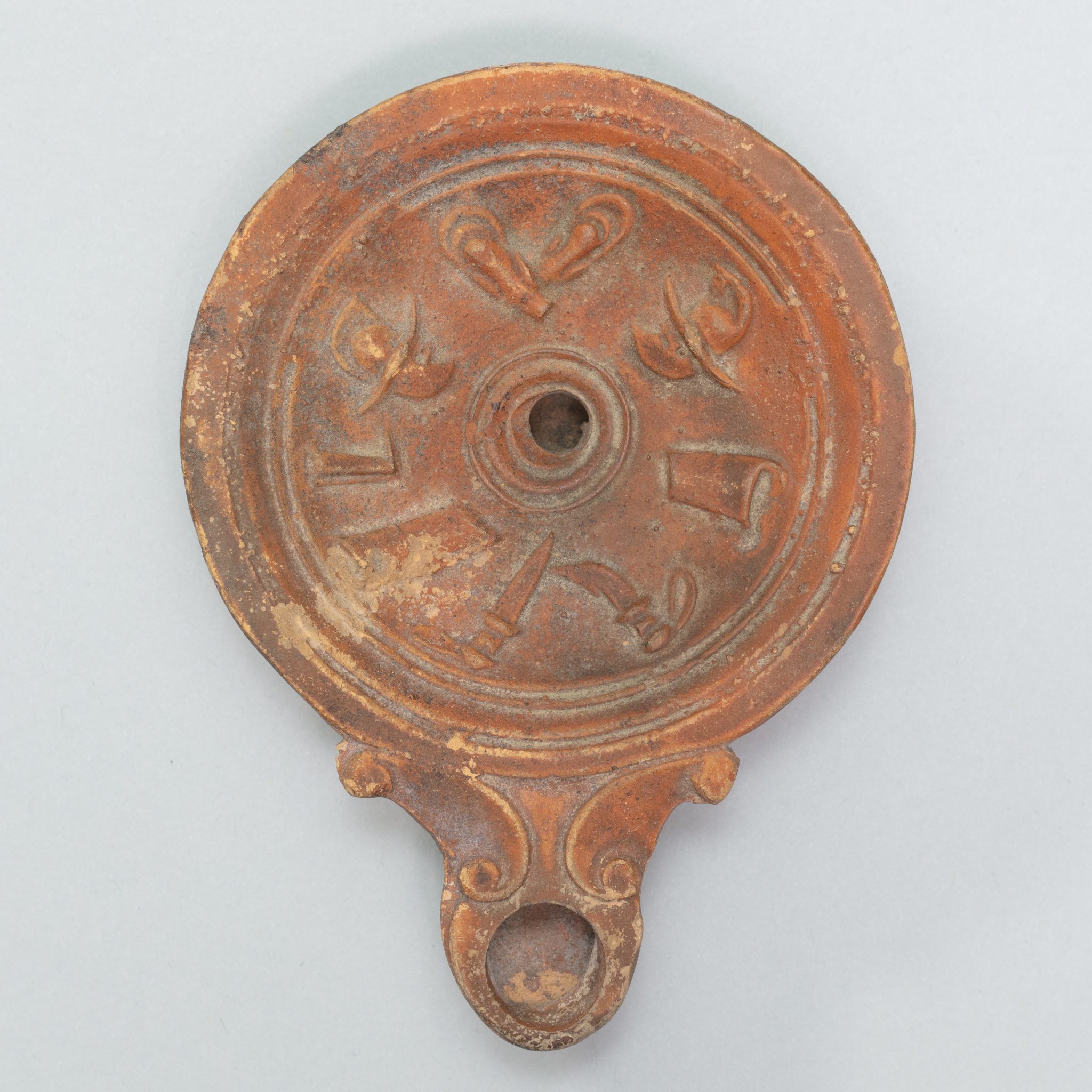 Circular terracotta lamp with gladiatorial weapons carved in a circle on its surface.