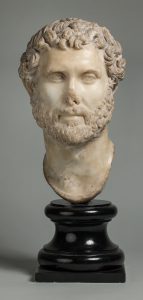 White marble portrait head of a man with short curly hair and a beard, mounted on a black base. The nose is partially broken off.
