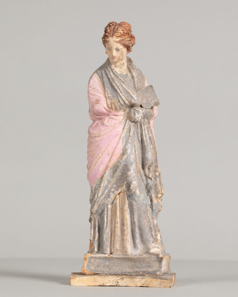 Painted terracotta statue of a woman with red hair, gather her pink and gray robe into her arms.