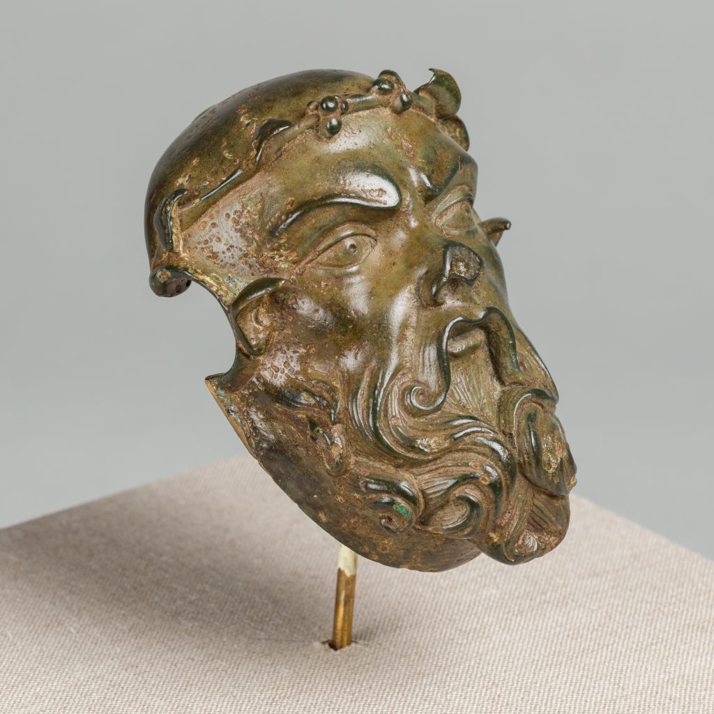 Bronze mask of a man's face with a curly beard and an ivory wreath, mounted on a post.