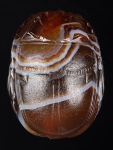 Alternate view of Onyx, scarab-shaped cameo depicting Poseidon pulling his trident from a rock. A white stripe curves across the amber-colored surface.
