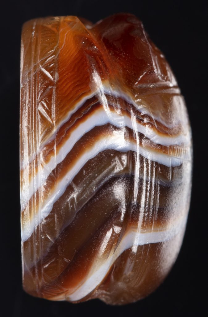 Alternate view of Onyx, scarab-shaped cameo depicting Poseidon pulling his trident from a rock. A white stripe curves across the amber-colored surface.
