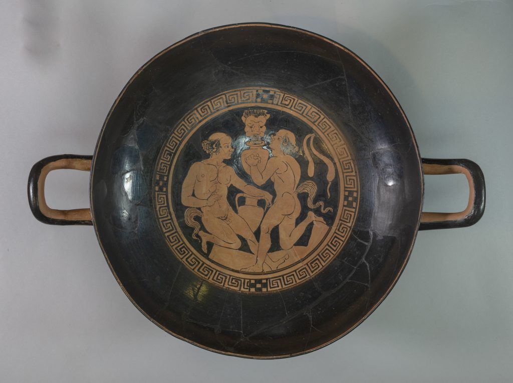 Birds-eye view of a black terracotta cup with two handles. The center features a circular, red-figure painting of three figures surrounded by a scroll pattern.
