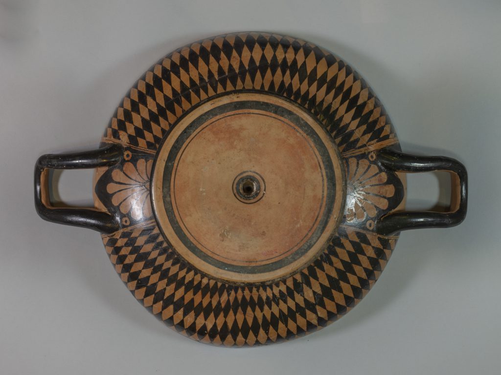 Alternate view of Birds-eye view of a black terracotta cup with two handles. The center features a circular, red-figure painting of three figures surrounded by a scroll pattern.
