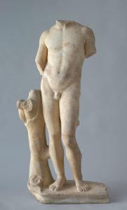 Alternate view of a white marble status of a young man's body standing against the base of a tree. The arms and head have broken off and are not present.