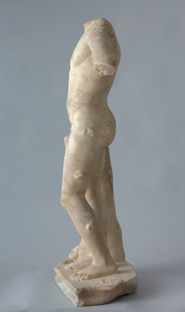 Alternate view of a white marble status of a young man's body standing against the base of a tree. The arms and head have broken off and are not present.