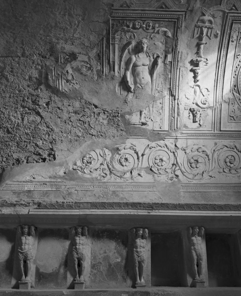 Black and white photograph with a detail of stone figural carvings in the curved wall of a bath.