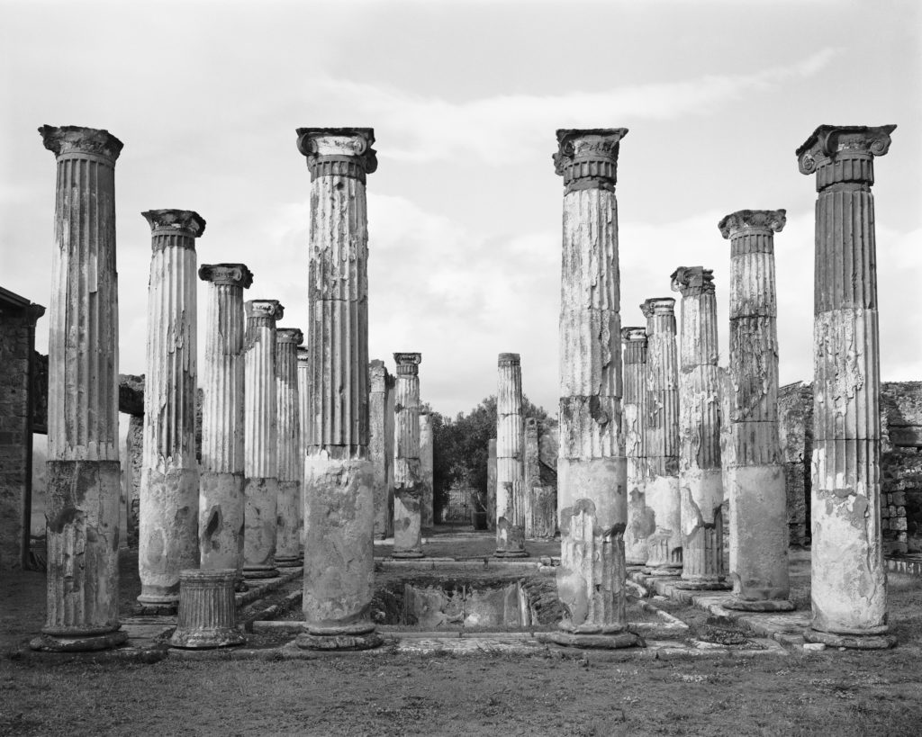 Black and white photograph featuring a head-on view of four rows of capital columns extending into the distance.