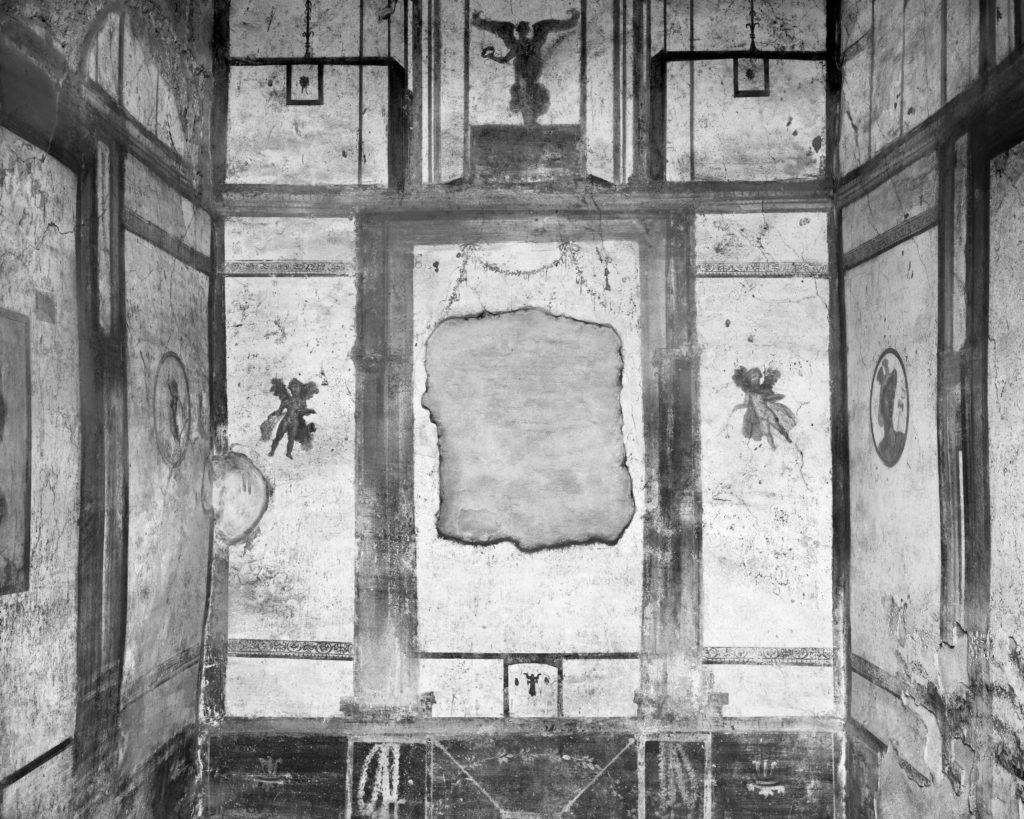 Black and white photo of the ruins of a rectangular interior space with small winged figures painted in the wall panels.