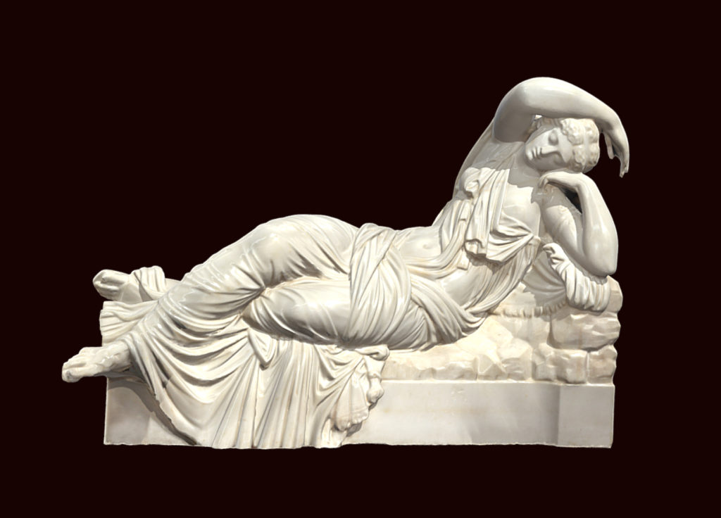 Alternate view of Statue of a reclined female wearing a toga, on display between marble columns. Statue sits on a block carved with warriers and snakes.