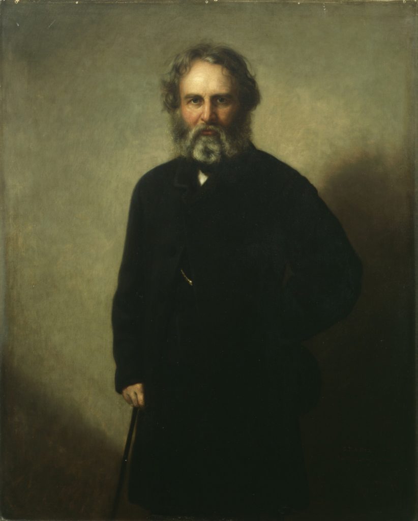 Shadowy portrait of a man with gray hair and a long beard, dressed in a long black suit jacket.
