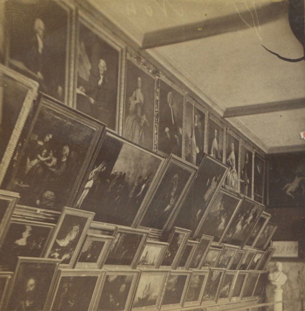 Black and white photo of a wall of framed artworks, hung side-by-side and extending up to the ceiling.