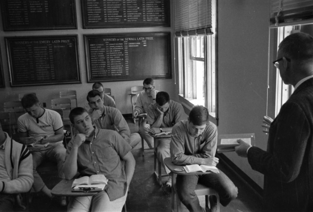 Photograph of a several young men at desks in a classroom, watching a professor lecture.