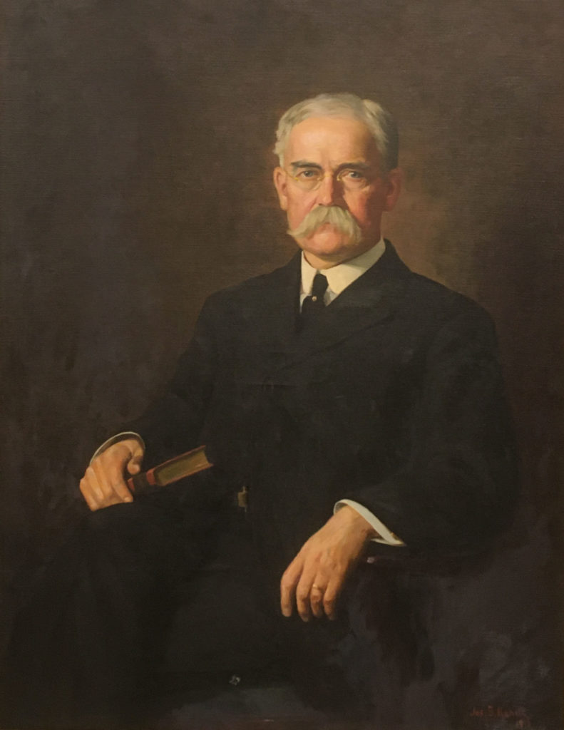 Portrait of a man with white hair, glasses, and a long mustache, seated in a chair and holding a book.