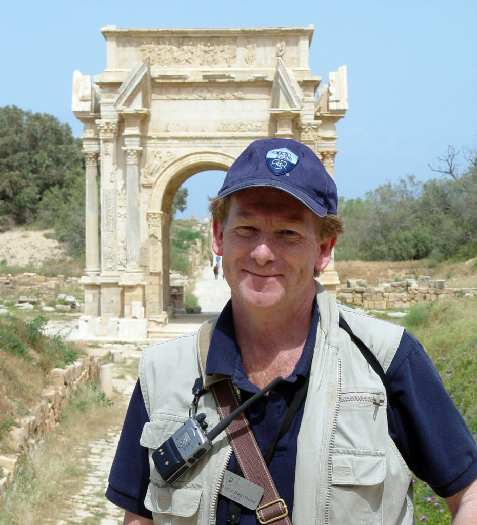 Photograph of a man wearing a cap and a safari-style vest, with the ruins of an arch behind him.