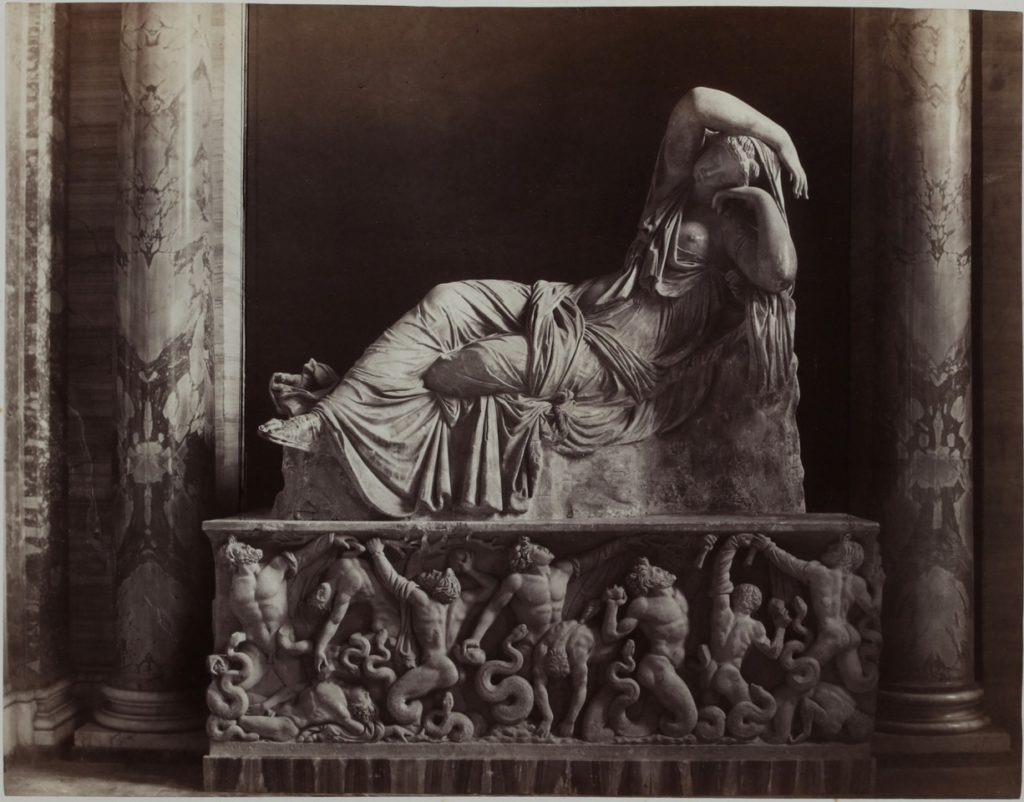 Statue of a reclined female wearing a toga, on display between marble columns. Statue sits on a block carved with warriers and snakes.